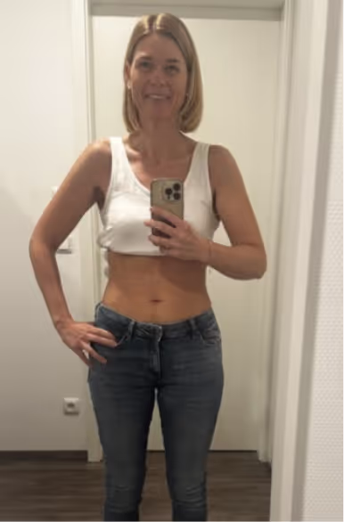 Eine lächelnde blonde Frau in Jeans und weißem Oberteil macht ein Selfie im Spiegel.