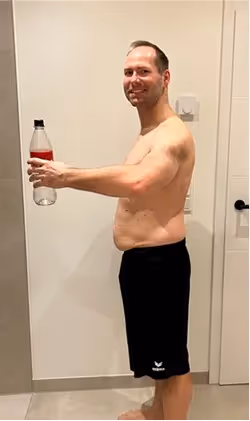 Ein lächelnder Mann ohne Hemd in Shorts hält eine Flasche vor sich.