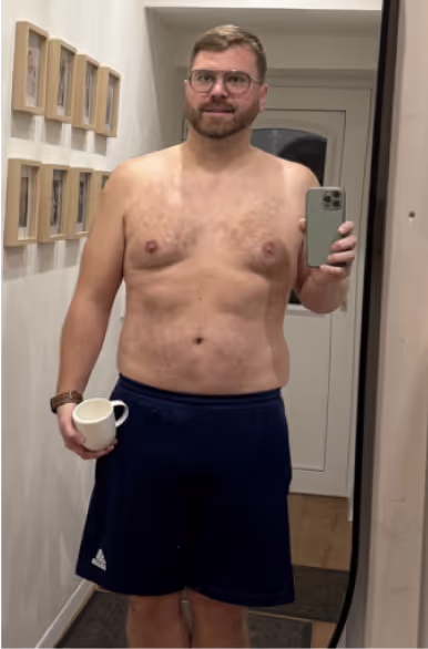 Ein lächelnder Mann ohne Hemd, in Shorts und mit Brille macht ein Selfie im Spiegel und hält eine Tasse.