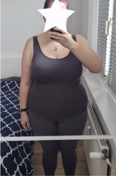 Eine Frau in einem Tanktop und Leggings macht ein Selfie im Spiegel, ihr Gesicht ist mit einem Stern-Emoji bedeckt.