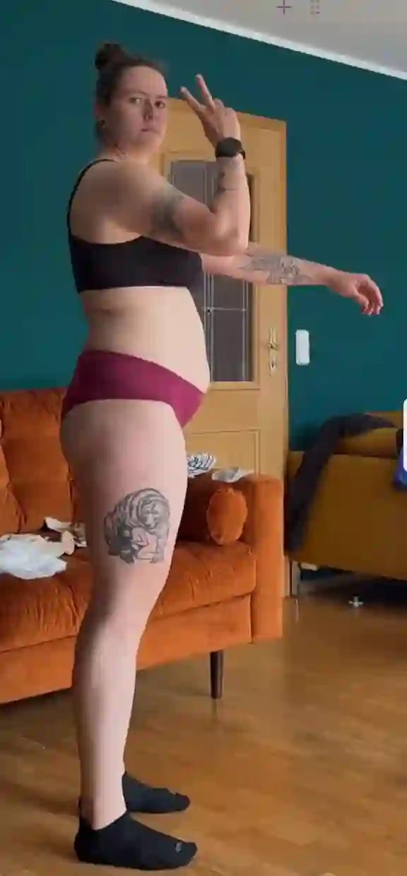 Eine Frau in Sport-BH und Unterwäsche zeigt das Friedenszeichen und steht seitlich vor einem Sofa.